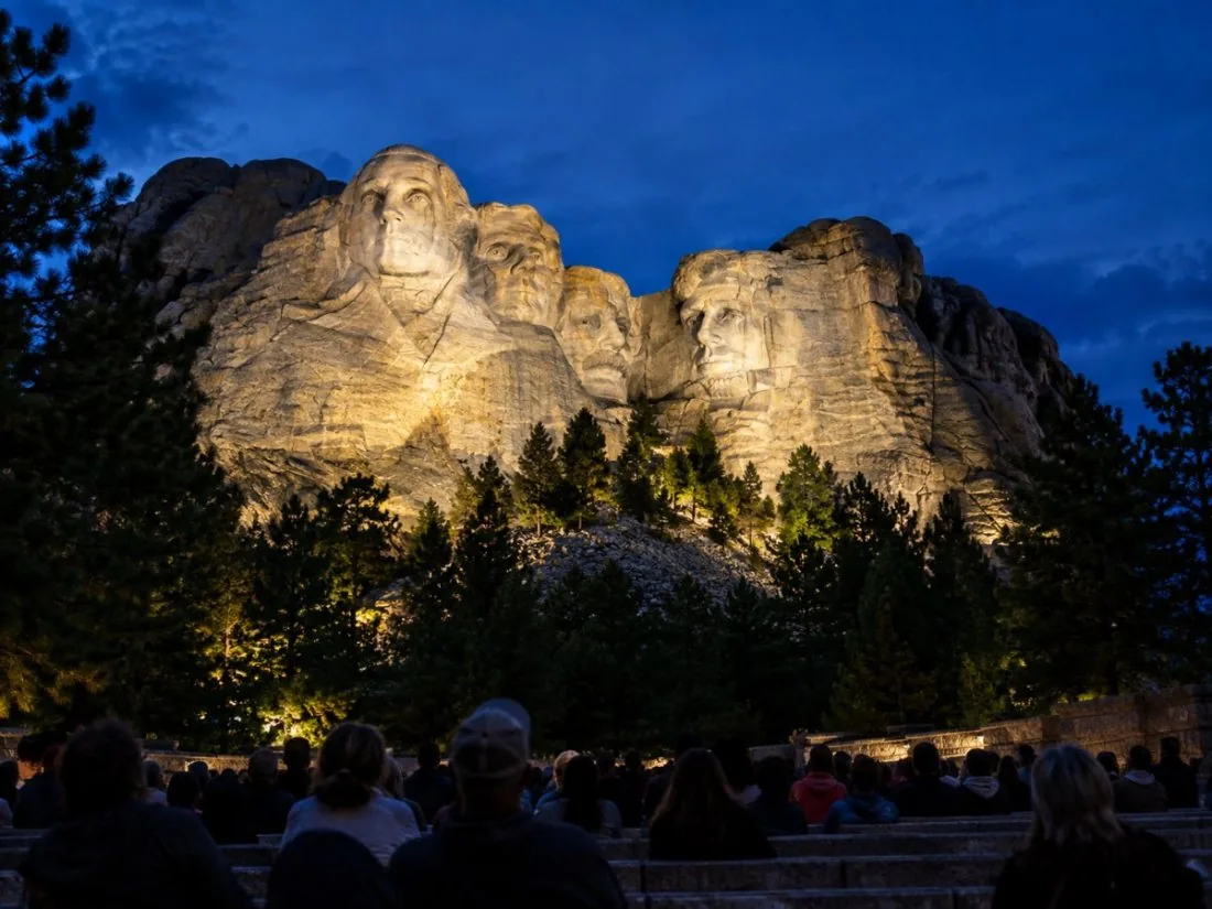 Mount Rushmore lit by floodlights at dusk above an outdoor amphitheater